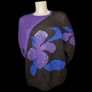 Può includere: Un maglione viola e grigio con un motivo floreale. Il maglione ha un girocollo e maniche lunghe. Il motivo floreale è nelle tonalità del viola e del blu, con accenti di paillettes bianche. Il maglione è esposto su un manichino.