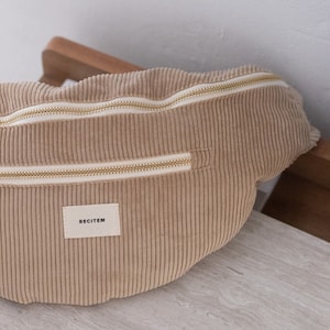 Peut inclure: Une banane en velours côtelé beige avec une fermeture éclair dorée et une étiquette blanche sur laquelle est inscrit "RECITEM".