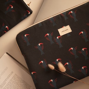 Peut inclure: Une housse d'ordinateur portable noire avec une fermeture éclair et un motif répétitif de toucans sur un fond foncé. La housse porte une étiquette blanche avec le texte "atelier bobbie".