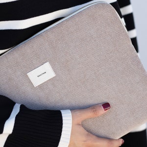 Könnte beinhalten: Eine beige Laptop-Hülle mit Reißverschluss, gehalten von einer Person, die einen schwarz-weiß gestreiften Pullover trägt. Ein kleines weißes Etikett mit dem Wort "RECITEM" ist an der Hülle befestigt.