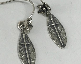May include: Silver earrings with a cross design and a flower detail. The cross is on a textured oval shape.