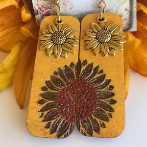 May include: A pair of gold-toned sunflower earrings with a wood base. The earrings feature a large sunflower design with a brown and red center and gold glitter accents. The earrings are attached to gold hooks.