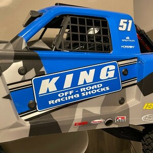 May include: A blue and grey radio-controlled truck with a black roll cage and a "KING OFF-ROAD RACING SHOCKS" sticker. The truck has a number "51" on the side and the words "BAJA" and "ISP" on the back.