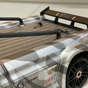 May include: A grey and black remote control truck with a camouflage paint job. The truck has a black roll cage, a black rear wing, and a "Horizon" sticker on the side. The truck is sitting on a grey surface.