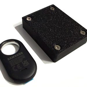 May include: Black Samsung device with a silver ring and a textured black rectangular box with four silver screws. The Samsung device has text and symbols, including "CE" and "UK CA".