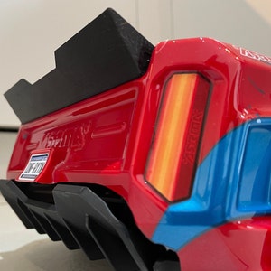 Puede incluir: Un camión de juguete rojo, azul y blanco con un alerón negro y un difusor trasero. El camión tiene un logotipo "V-Maxx" en el lateral y un logotipo "Monster" en la parte trasera.