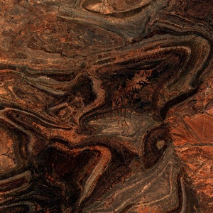 May include: An aerial view of a geological formation with layers of red, brown, and grey rock. The image shows a complex pattern of folds and curves, with a textured surface and a natural, earthy aesthetic.