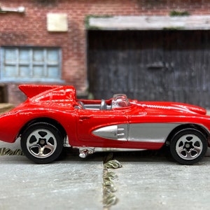 May include: A red toy car with silver accents and black wheels. The classic car has a streamlined design and is a miniature replica of a vintage vehicle. The toy car is a collectible item and is displayed on a gray surface.