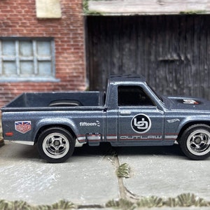 Könnte beinhalten: Ein graues Hot Wheels Outlaw Spielzeug-Pick-up mit Chromrädern. Der Truck hat rote und weiße Streifen, die Nummer 5 und die Schriftzüge "fifteen" und "Outlaw". Auf der Seite ist eine Union Jack-Flagge zu sehen.
