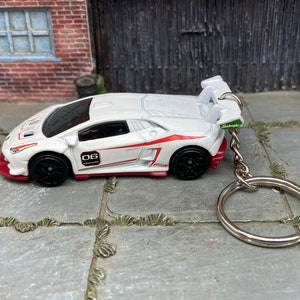 Op de afbeelding: Witte en rode Lamborghini sleutelhanger met zwarte wielen. De miniatuurauto heeft het nummer "06" op de zijkant en een achterspoiler. De sleutelhanger is bevestigd aan een zilveren metalen ring.
