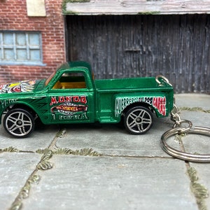Puede incluir: Un llavero de camioneta de juguete verde con ruedas negras y plateadas. El camión tiene las palabras "Mundo Hot Wheels" y "Guerrero De Dos Caras" impresas. El llavero es de metal.