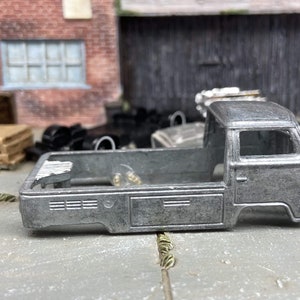 May include: A silver toy truck body with a flatbed design. The truck has a cab with a window and door detail. The background includes a brick wall, a wooden pallet, and other vehicle parts.