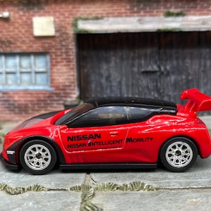 May include: A red and black toy car with the text "NISSAN INTELLIGENT MOBILITY" on the side. The car has a spoiler and silver wheels. The toy car is a miniature replica of a sports car.