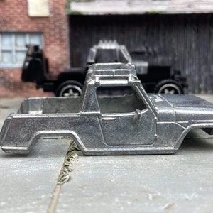 May include: A silver-colored toy truck body with a roll bar, a cab, and a bed. The truck is made of metal and appears to be a miniature replica. Another black toy truck is visible in the background.