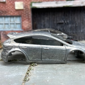 May include: A silver die-cast model car, a hatchback, with a spoiler. The car is missing its wheels and interior. The model is set on a concrete surface with a brick building in the background.