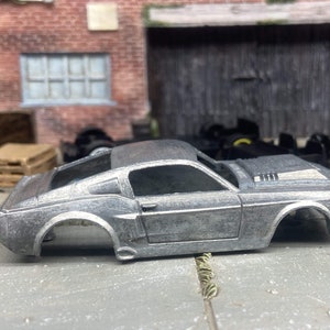 May include: A silver-colored model car, a classic muscle car, sits on a concrete surface. The car lacks wheels and has a detailed body, including a roof, doors, and hood. The background features a brick wall and a weathered wooden structure.