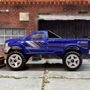 Può includere: Un camioncino giocattolo Ford F-150 blu con grandi pneumatici fuoristrada. Il camion ha strisce bianche e arancioni sul lato e la scritta "F-150" sul retro. Il giocattolo è ambientato su uno sfondo di muro di mattoni.