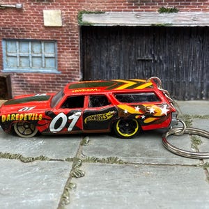 May include: A red die-cast station wagon toy car keychain featuring black flame graphics, the number 01 in white, the word DAREDEVILS in yellow, and the Hot Wheels logo. The vehicle has black wheels with yellow rims and a silver metal keyring attached.