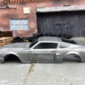May include: A silver-colored model car body, a classic muscle car design, sits on a concrete surface. The car lacks wheels and interior components. The background features a brick building and a wooden door.