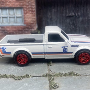 Könnte beinhalten: Ein weißes Hot Wheels Datsun Pickup-Truck-Spielzeug mit roten Rädern und blau-orangen Streifen. Der Spielzeug-Truck hat das Hot Wheels-Logo und die Nummer 71 auf der Seite. Der Truck steht auf einer grauen Oberfläche.
