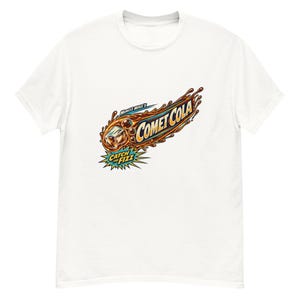 May include: A white short-sleeve t-shirt featuring a graphic of a comet made of cola liquid and an ice cube. The text reads "Muncle Mike's COMET COLA" and "CATCH THE FIZZ" in a stylized, colorful font with orange, brown, and yellow accents.