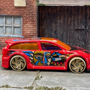 Puede incluir: Un coche de juguete Hot Wheels rojo con un diseño único. El coche presenta un diseño de rostro abstracto colorido en el lateral, ruedas doradas y el logotipo de Hot Wheels. El coche tiene un alerón y cristales tintados.
