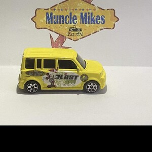 May include: A yellow toy car with black wheels and the words "BLAST OFF!" and a Mickey Mouse graphic on the side. The car has a surfboard design and the Disney logo. The car is a miniature replica.