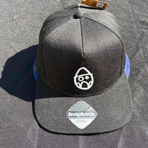 May include: A black snapback cap featuring a white embroidered graphic of a stylised egg-shaped character wearing a hat on the front panel. The flat brim includes a clear sticker with the text Snapback Original Headwear and a brown paper tag.