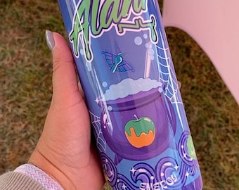 May include: A cylindrical tumbler with a blue background and the text "Witch's Brew" and "Alani" in green and white. The design includes a purple cauldron with an apple and a butterfly. The tumbler is flavored with other natural flavors.