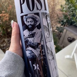 May include: A white tumbler featuring black and white images of a musician. The word "POST" is printed in bold, black letters at the top. The tumbler is held by a person.