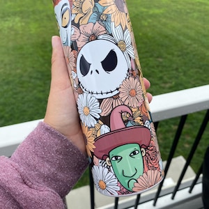 Op de afbeelding: Een witte tumbler met een bloemenontwerp met de personages Jack Skellington, Oogie Boogie en Sally uit de film The Nightmare Before Christmas.
