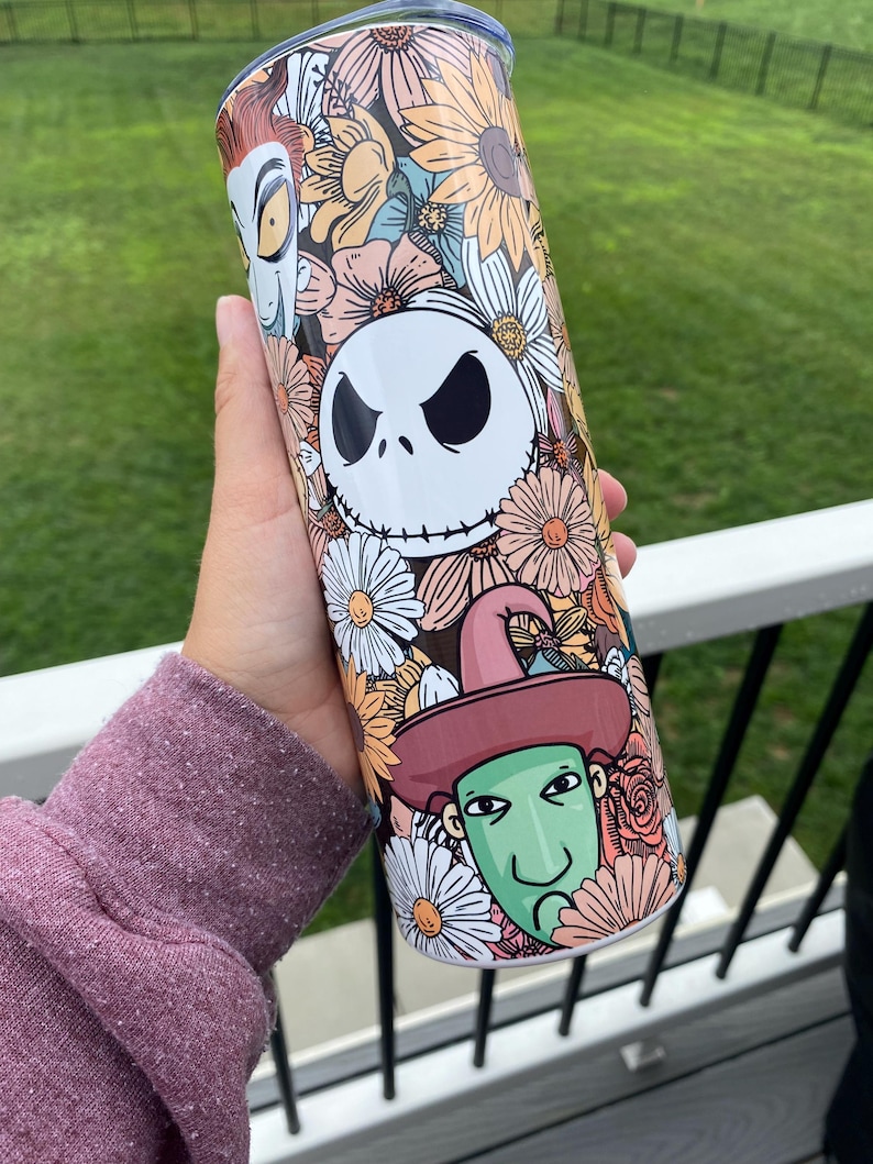 Op de afbeelding: Een witte tumbler met een bloemenontwerp met de personages Jack Skellington, Oogie Boogie en Sally uit de film The Nightmare Before Christmas.