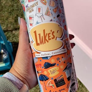 May include: A stainless steel tumbler featuring a colorful design with the text "Luke's" and "Stars Hollow, Connecticut." The tumbler has various illustrations, including pizza slices, coffee cups, and a film clapper.