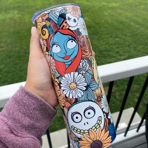 Op de afbeelding: Een roestvrijstalen beker met een kleurrijk bloemenontwerp met Jack Skellington en Sally uit The Nightmare Before Christmas.