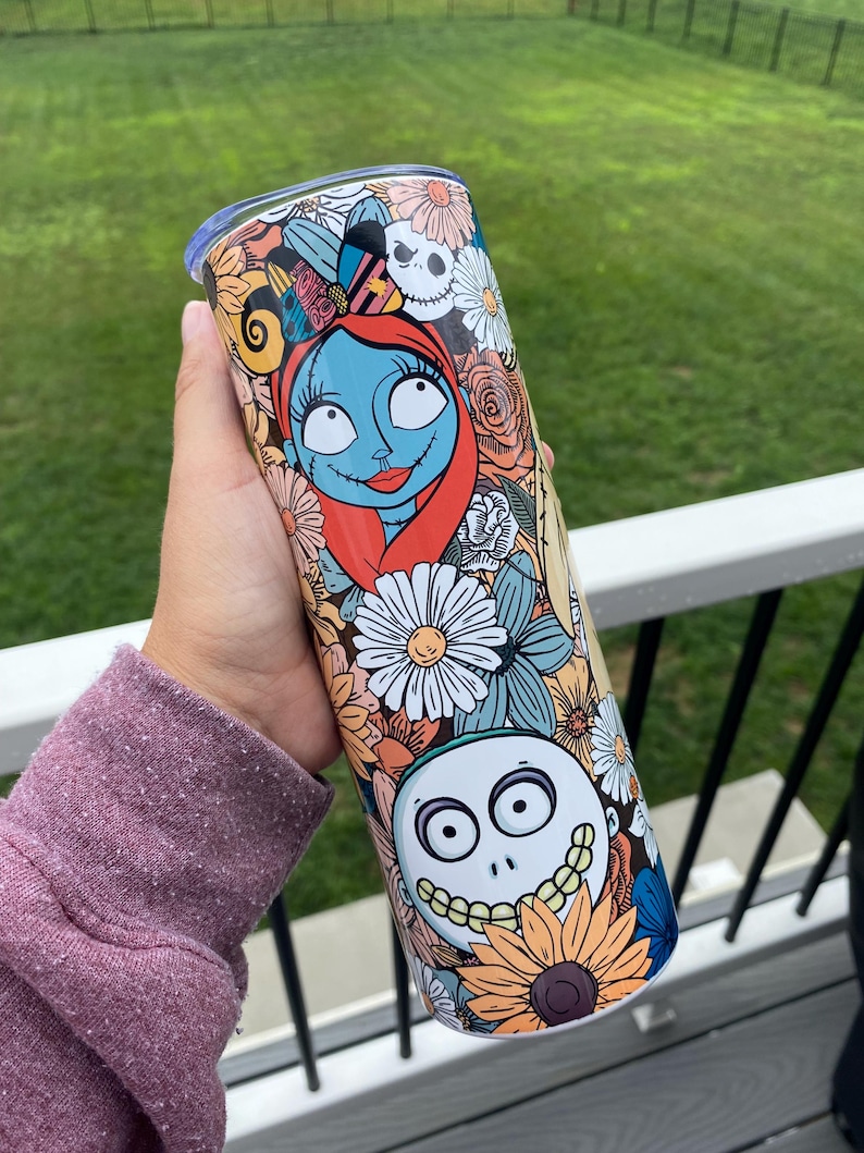 Op de afbeelding: Een roestvrijstalen beker met een kleurrijk bloemenontwerp met Jack Skellington en Sally uit The Nightmare Before Christmas.