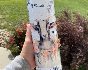 May include: A white stainless steel tumbler with a floral design and a cute goat face. The goat has black and white fur and is surrounded by pink and peach flowers.