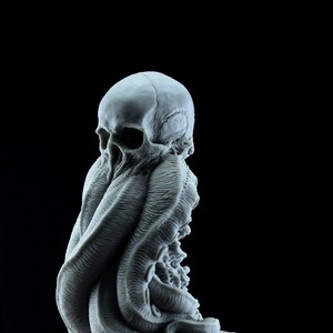 May include: A gray 3D printed sculpture of a skull with tentacles emerging from its head. The sculpture is on a base that resembles a rocky surface.