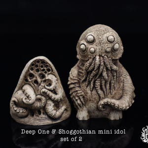 Puede incluir: Un conjunto de dos pequeñas esculturas de resina blanca detalladas. Una escultura es un Deep One, una criatura humanoide con tentáculos y una cabeza de pez. La otra escultura es un Shoggoth, una criatura con forma de blob con múltiples ojos y tentáculos. Las esculturas están etiquetadas "Deep One & Shoggoth mini idol set of 2".