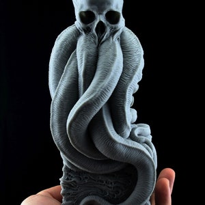 May include: A gray 3D printed sculpture of a creature with a skull-like head and tentacles. The creature is sitting on a base with a textured surface.