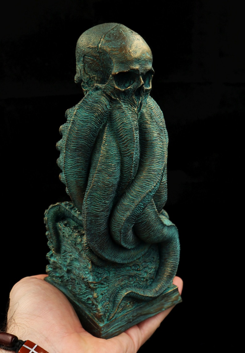 May include: A green and bronze colored statue of a skull with tentacles. The statue is on a square base and is being held in a person's hand.