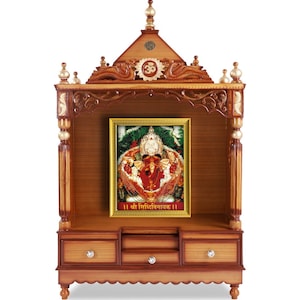 May include: A wooden temple with a framed image of a deity. The temple is made of brown wood with decorative carvings and gold accents. The image is in a gold frame and features a colorful depiction of a Hindu deity. The temple has drawers and a decorative top.