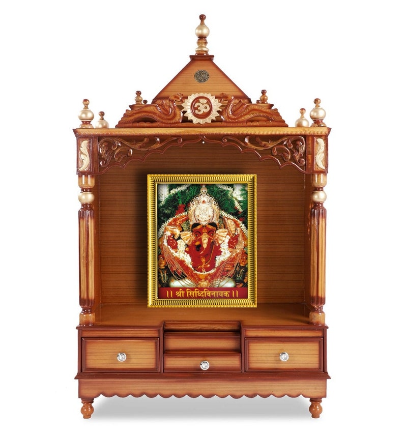 May include: A wooden temple with a framed image of a deity. The temple is made of brown wood with decorative carvings and gold accents. The image is in a gold frame and features a colorful depiction of a Hindu deity. The temple has drawers and a decorative top.