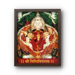 May include: Framed image of a red deity with a golden crown and jewelry, surrounded by garlands and greenery. The image has text in a foreign language at the bottom and is set in a dark brown frame.