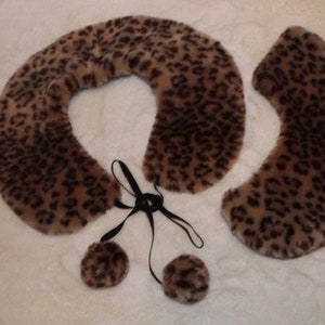 May include: A brown and black faux fur collar with a leopard print pattern. The collar has a black ribbon tie and two brown and black faux fur pom-poms.