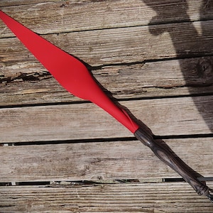 Peut inclure: Une lance rouge avec un manche en bois foncé. La pointe de la lance est d'un rouge vif, passant à un manche en bois foncé et texturé. La poignée a une boucle à l'extrémité. La lance est posée sur une surface en bois.