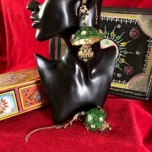May include: A pair of green and gold earrings with a dangling design. The earrings are on a black mannequin head. The earrings have a green and gold design with a round shape and a dangling design.