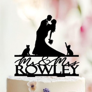 May include: A two-tiered wedding cake with floral decorations and a topper. The topper is a black silhouette of a kissing couple, two cats, and the words "Mr. & Mrs. Rowley". The cake is decorated with purple, yellow, and white flowers.