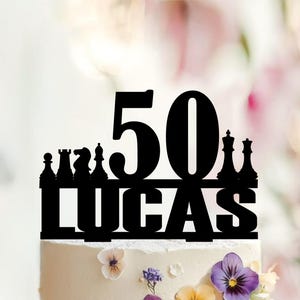 Puede incluir: Un adorno para pastel en color negro que muestra el número 50 y el nombre LUCAS en letras mayúsculas negritas. El diseño incorpora siluetas de varias piezas de ajedrez, incluyendo peón, torre, caballo, alfil, rey y reina, en una línea.