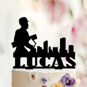 Op de afbeelding: Een zwarte taarttopper met een silhouet van een bouwvakker die blauwdrukken vasthoudt naast een stadssilhouet. De naam LUCAS staat in dikke hoofdletters aan de basis van het ontwerp, dat bovenop een wit geglazuurde taart is geplaatst.
