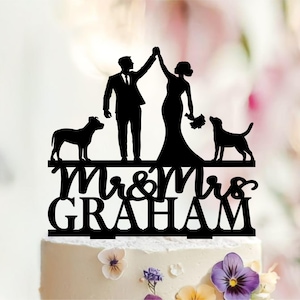 May include: A black silhouette cake topper featuring a couple standing with arms raised, flanked by two dogs. Below the figures, the text reads Mr & Mrs GRAHAM in a combination of cursive and block lettering, all mounted on a white frosted cake.